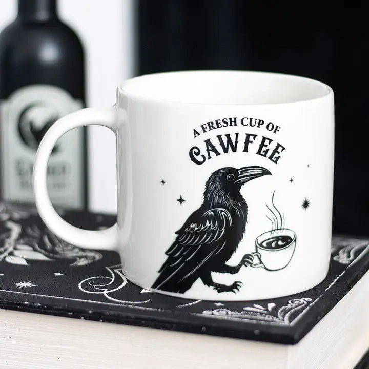 White mug with a black crow holding a coffee cup and text 'A Fresh Cup of Cawfee' on a dark coaster.