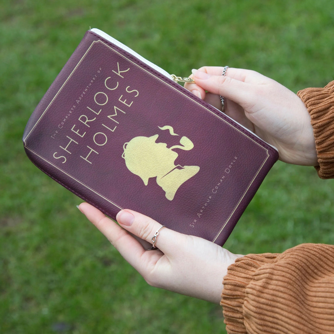 Hands holding out a Burgundy Zipped Pouch featuring a silhouette of Sherlock Holmes with gras in the background