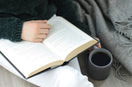 A person reading an open book with a dark mug beside them, wrapped in a cosy grey throw