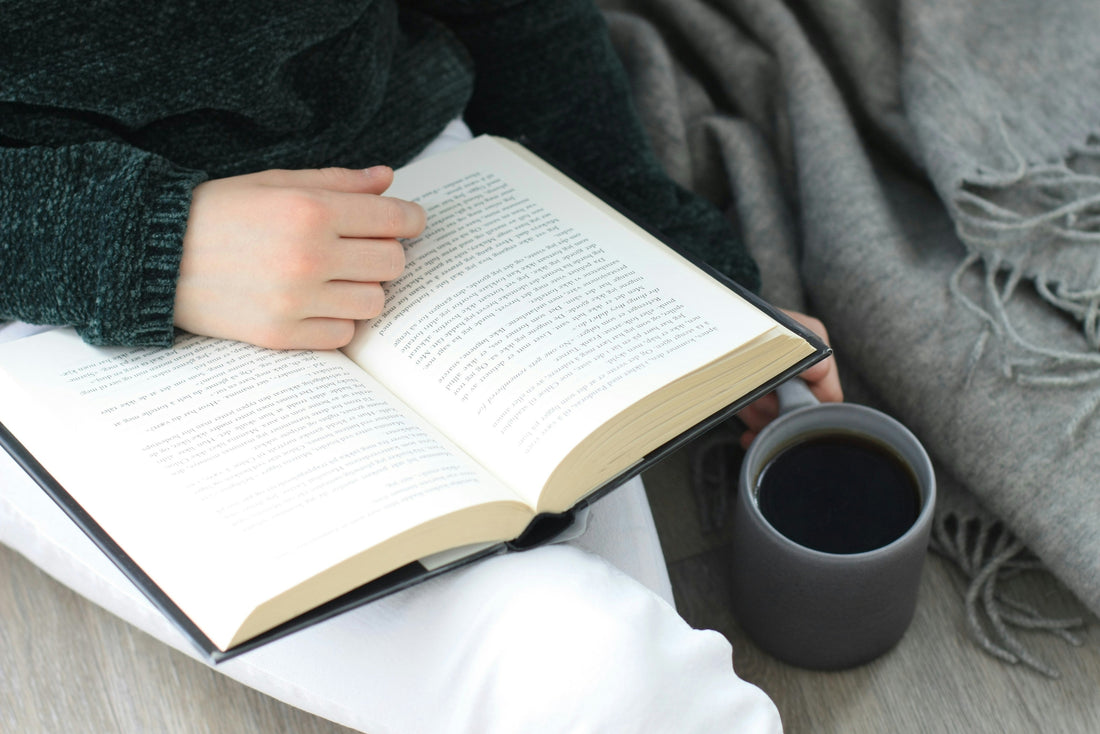 A person reading an open book with a dark mug beside them, wrapped in a cosy grey throw