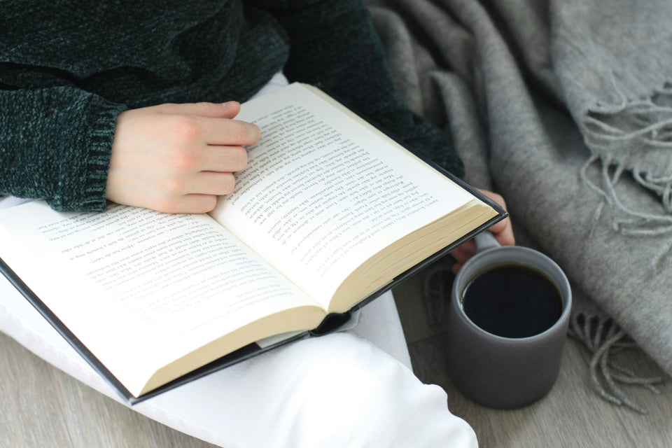 A person reading an open book with a dark mug beside them, wrapped in a cosy grey throw