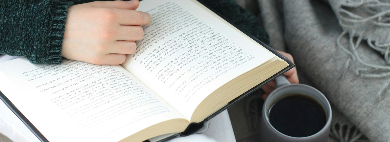 A person reading an open book with a dark mug beside them, wrapped in a cosy grey throw
