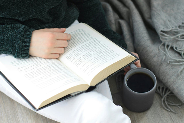 A person reading an open book with a dark mug beside them, wrapped in a cosy grey throw