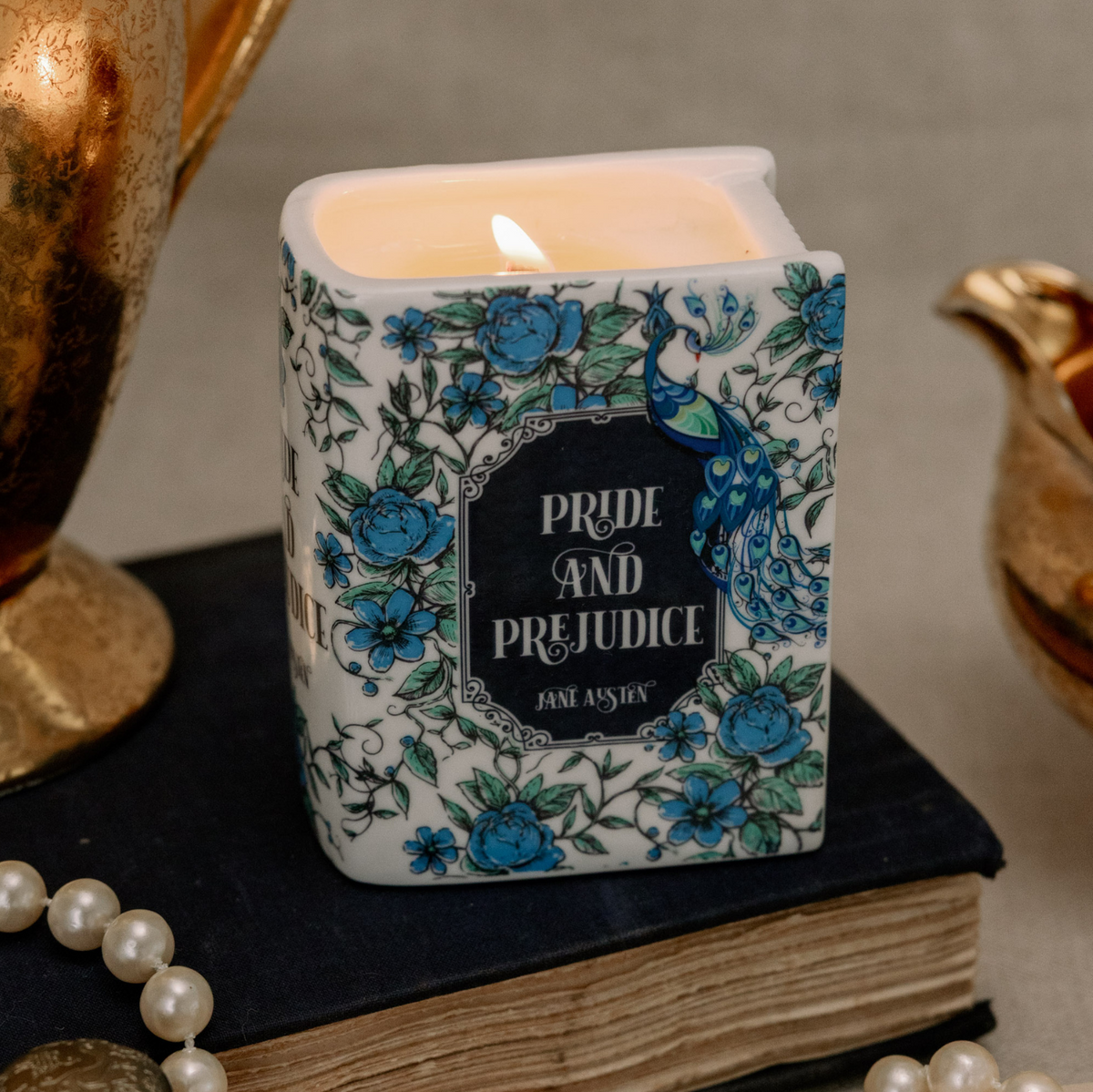 A square white candle with a blue floral and peacock pattern, lit and resting on a dark book. The front label reads 'PRIDE AND PREJUDICE' with 'JAM ASHE' below.