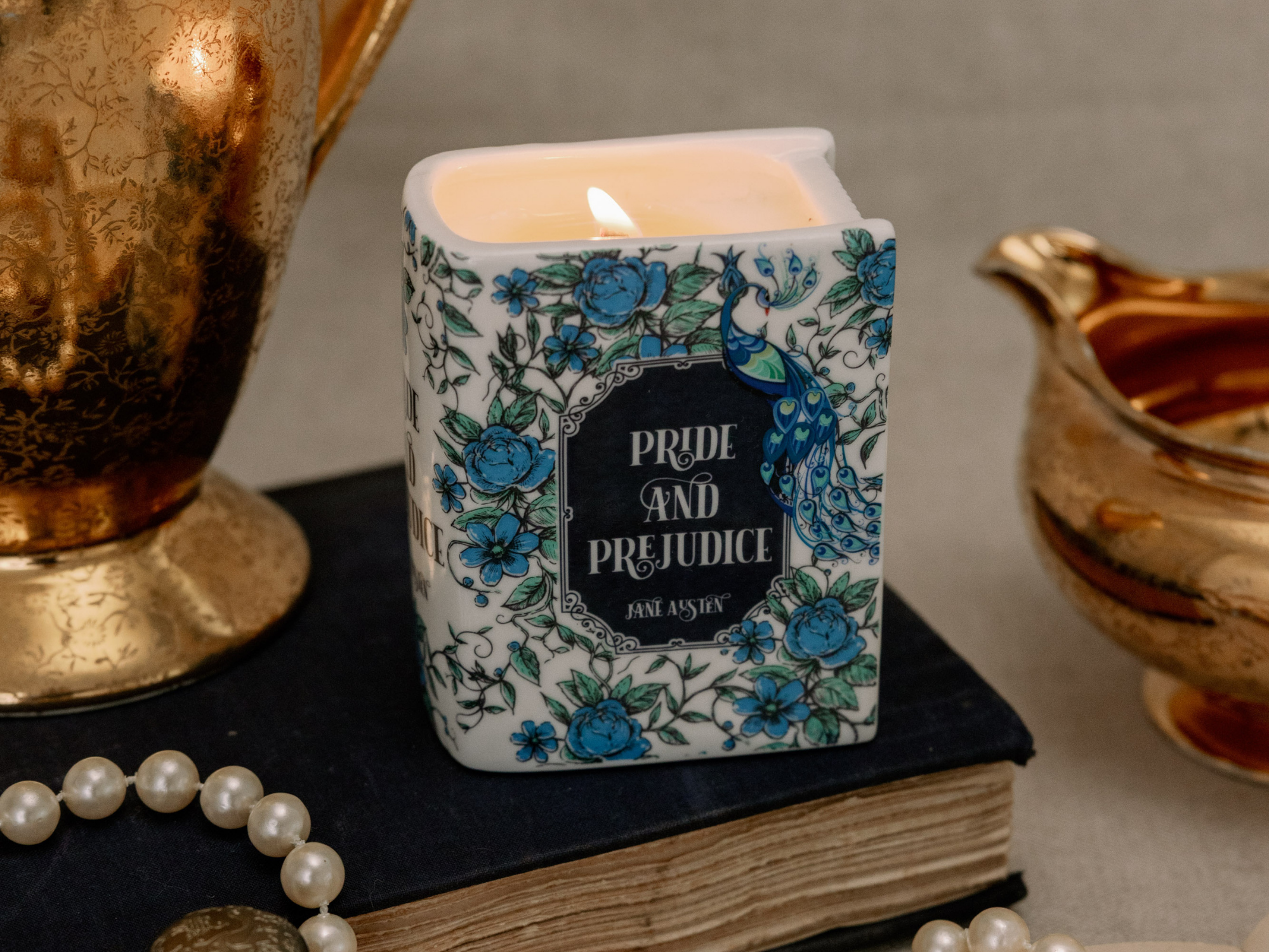 A square white candle with a blue floral and peacock pattern, lit and resting on a dark book. The front label reads 'PRIDE AND PREJUDICE' with 'JAM ASHE' below.