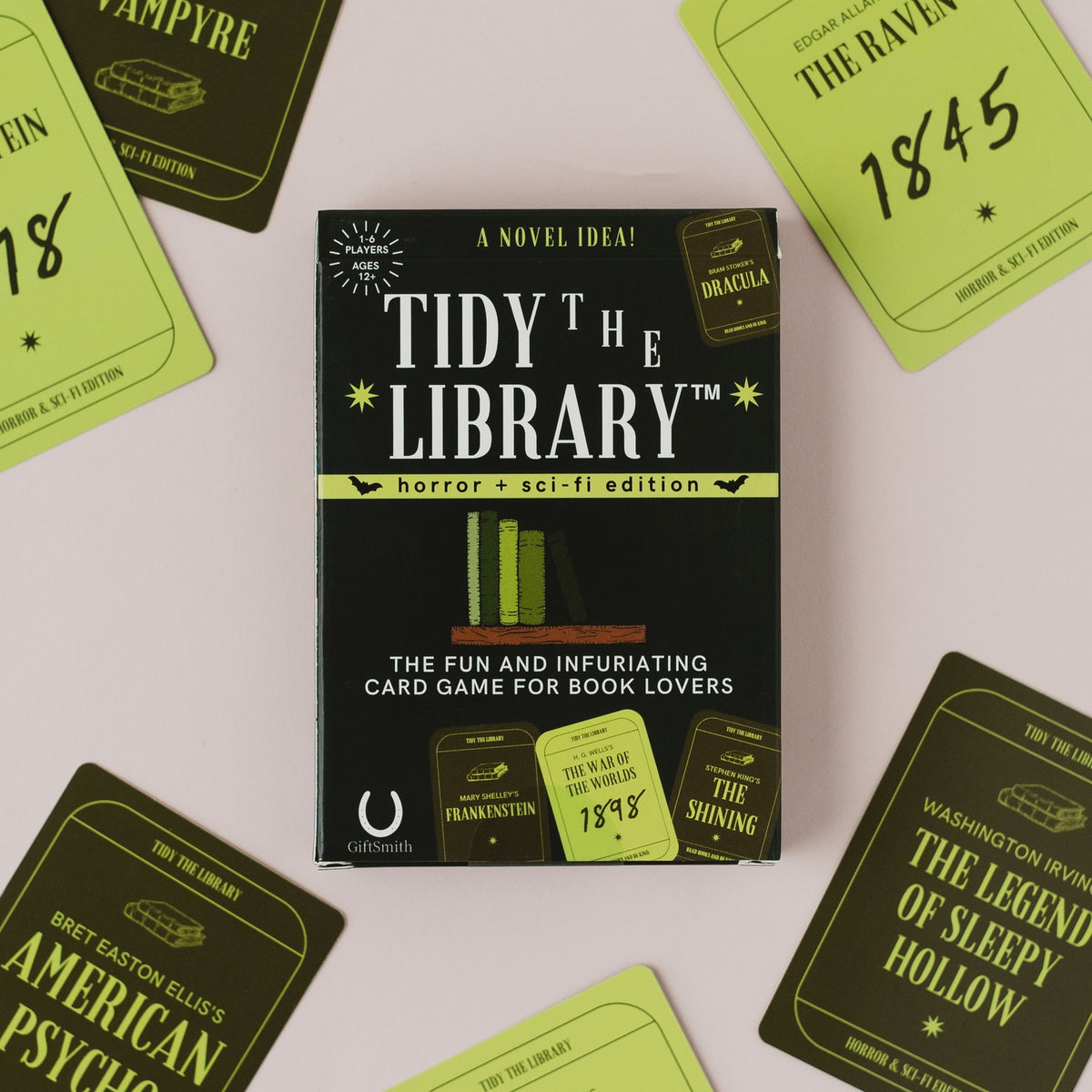 The 'Tidy the Library: Horror + Sci-Fi Edition' card game box is centered on a light pink background, surrounded by various themed card faces featuring titles like 'The Raven', '1845', 'Frankenstein', and 'The Shining'.