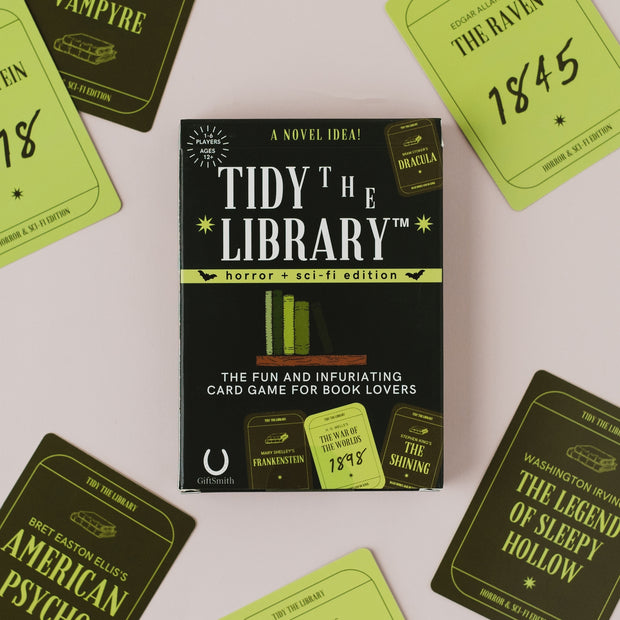 The 'Tidy the Library: Horror + Sci-Fi Edition' card game box is centered on a light pink background, surrounded by various themed card faces featuring titles like 'The Raven', '1845', 'Frankenstein', and 'The Shining'.