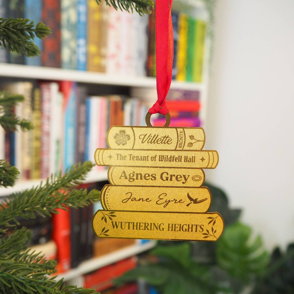 A brass ornament shaped like a stack of books, hanging from a red ribbon. The books are engraved with names and book titles, including 'Villette', 'The Tenant of Wildfell Hall', 'Agnes Grey', 'Jane Eyre', and 'Wuthering Heights'.