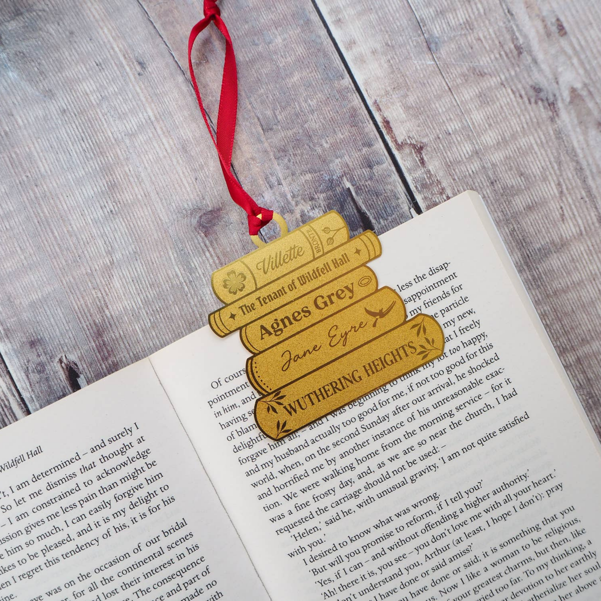 A brass ornament shaped like a stack of books, with a red ribbon resting on an open book with a grey wooden background. The books are engraved with names and book titles, including 'Villette', 'The Tenant of Wildfell Hall', 'Agnes Grey', 'Jane Eyre', and 'Wuthering Heights'.