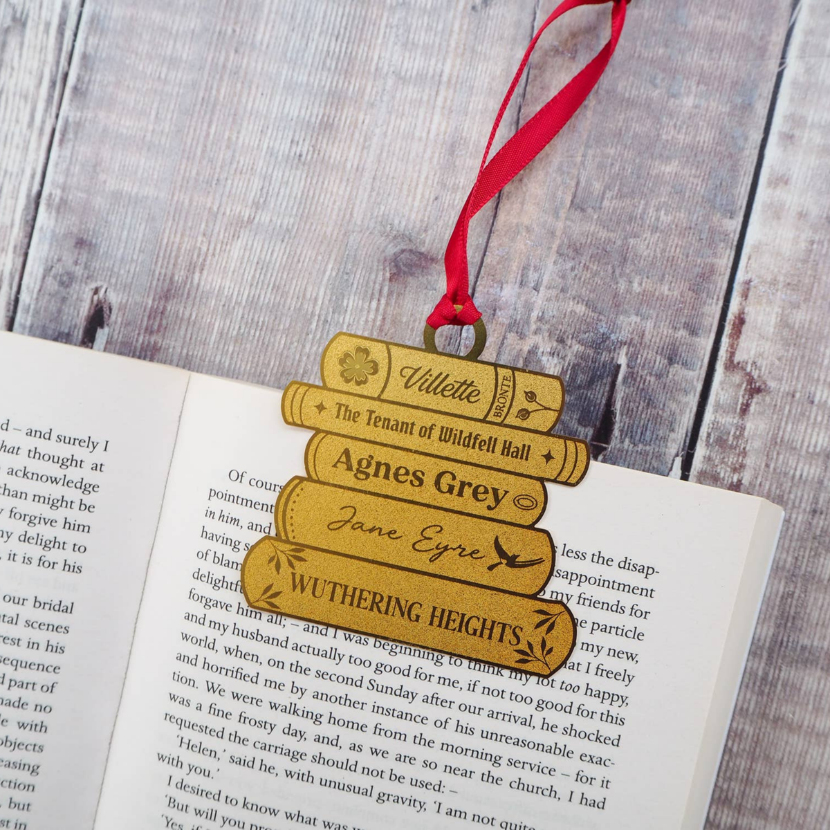 A brass ornament shaped like a stack of books, with a red ribbon resting on an open book with a grey wooden background. The books are engraved with names and book titles, including 'Villette', 'The Tenant of Wildfell Hall', 'Agnes Grey', 'Jane Eyre', and 'Wuthering Heights'.
