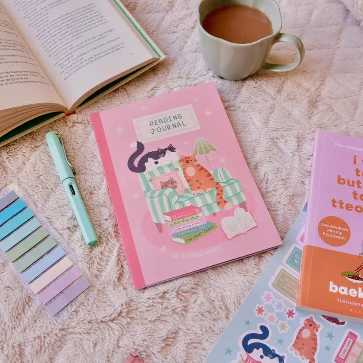 Pink reading journal, open book, mug of tea, and stationery items on a textured surface.