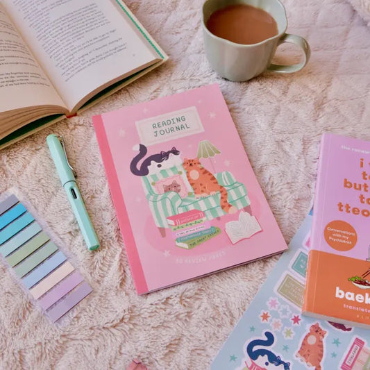Pink reading journal, open book, mug of tea, and stationery items on a textured surface.