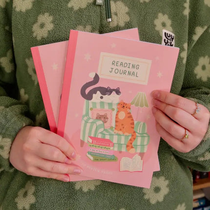Person holding a pink 'Reading Journal' with cat illustration, wearing a green floral-patterned garment.