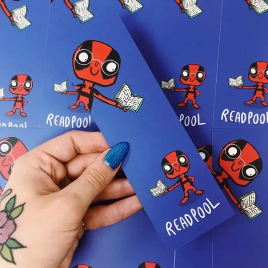 Hand holding a blue bookmark with a cartoon Deadpool character on a blue background with more bookmarks.