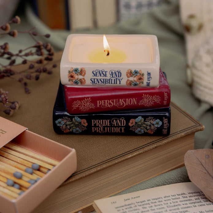 Stack of books with a lit candle on top, resembling Jane Austen's novels, next to matches and an open book.