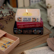 Stack of books with a lit candle on top, resembling Jane Austen's novels, next to matches and an open book.