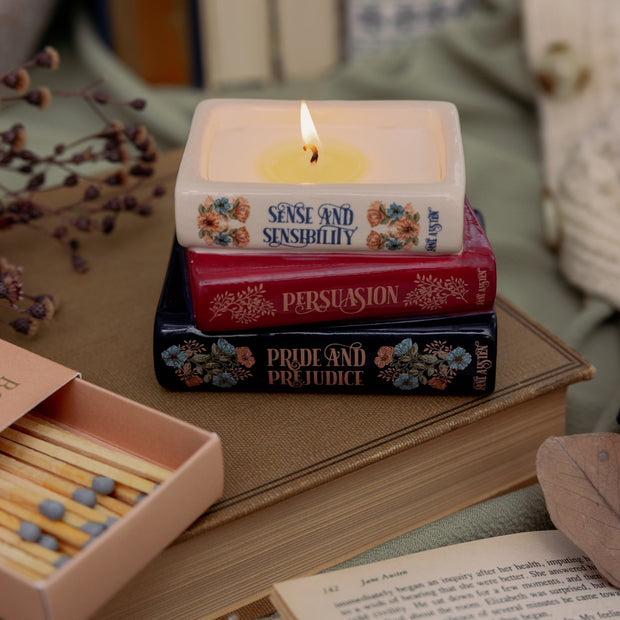 Stack of books with a lit candle on top, resembling Jane Austen's novels, next to matches and an open book.