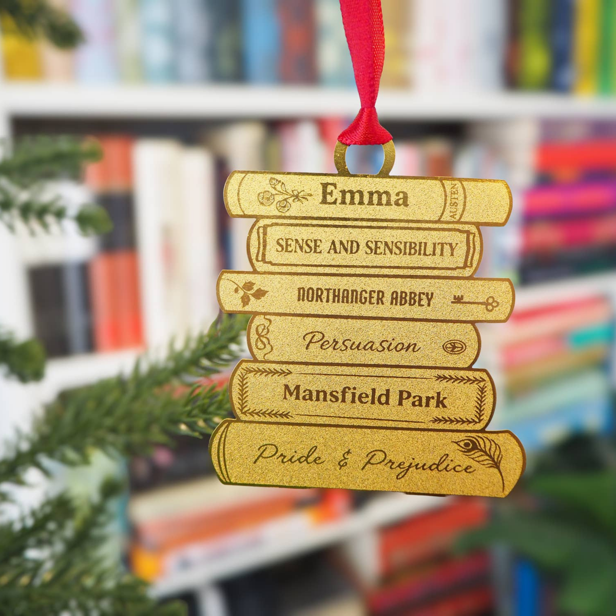 A gold-colored Christmas ornament shaped like a stack of books, hanging from a red ribbon. The books are engraved with names and phrases including 'Emma', 'Sense and Sensibility', 'Northanger Abbey', 'Persuasion', 'Mansfield Park', and 'Pride & Prejudice'.