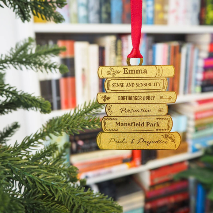 A brass book-stack Christmas ornament hanging from a red ribbon, featuring names and book titles from Pride and Juvenile, set against a blurred background of a bookshelf and green tree branches.