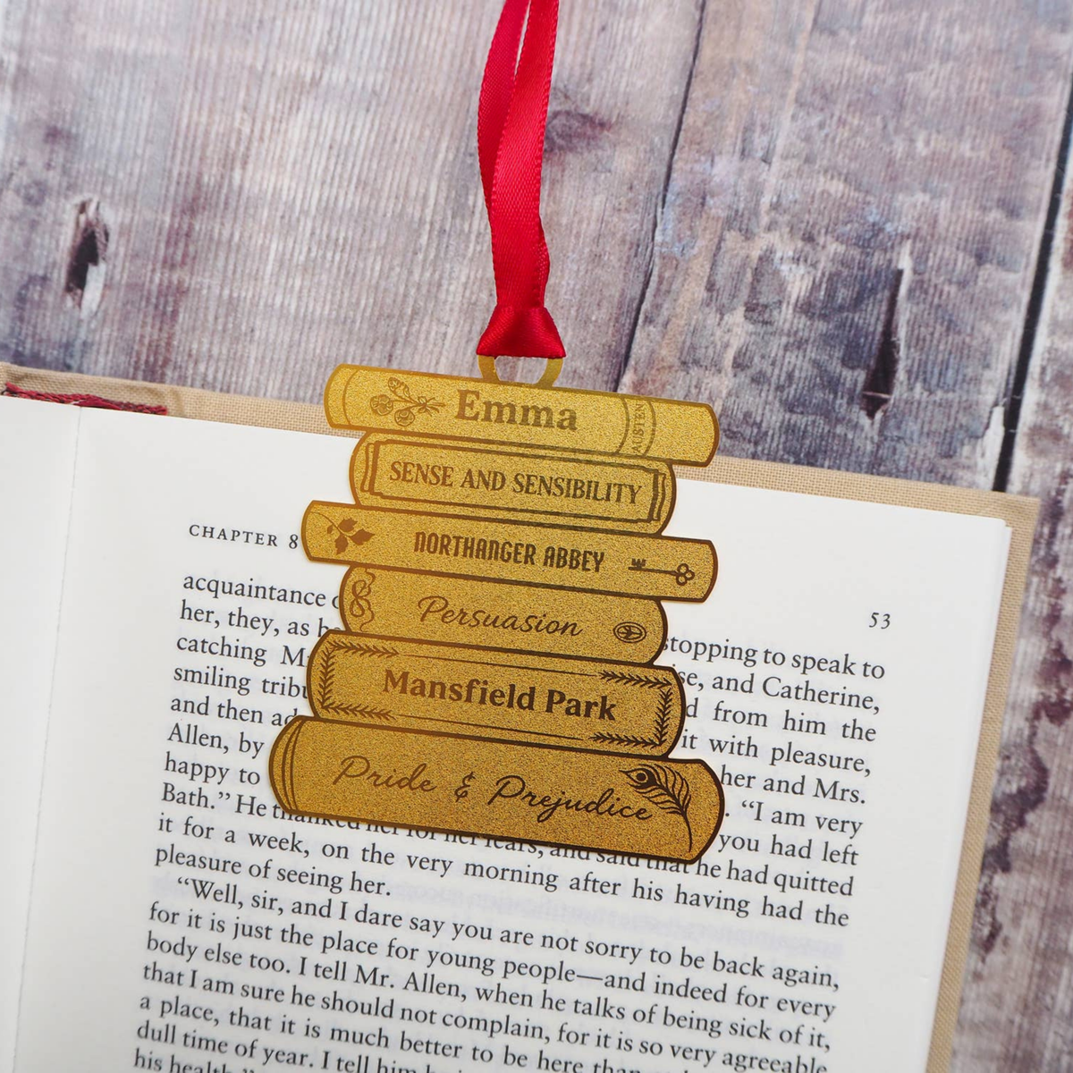 A stack of five brass books in a bookmark engraved with famous literary titles and names like Emma, Sense and Sensibility, Northanger Abbey, Persuasion, Mansfield Park, and Pride & Prejudice, hanging from a red ribbon on an open book.