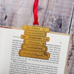 A stack of five brass books in a bookmark engraved with famous literary titles and names like Emma, Sense and Sensibility, Northanger Abbey, Persuasion, Mansfield Park, and Pride & Prejudice, hanging from a red ribbon on an open book.