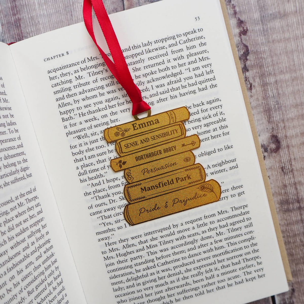 A close-up of an open book page with a red ribbon bookmark threaded through a brass bookstack bookmark labeled Emma, Sense and Sensibility, Pertinence Critique, and Pride and Prejudice, placed on a grey wood surface.