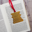 A close-up of an open book page with a red ribbon bookmark threaded through a brass bookstack bookmark labeled Emma, Sense and Sensibility, Pertinence Critique, and Pride and Prejudice, placed on a grey wood surface.