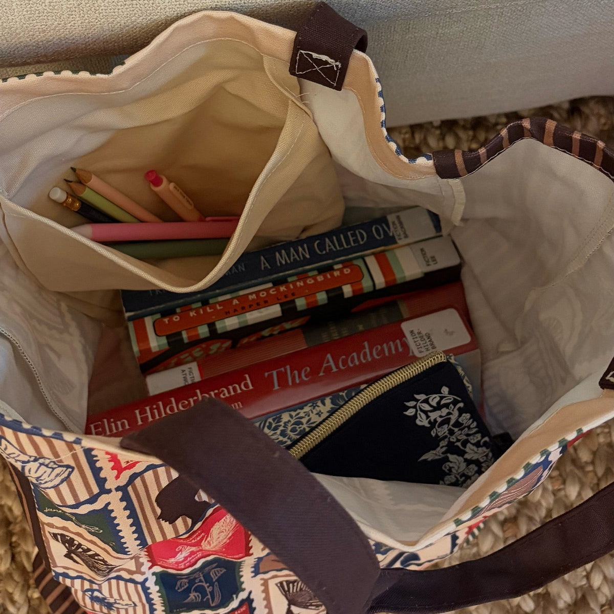 A close-up overhead view of a canvas tote bag filled with books, pencils, and a small zip pouch. The tote has a cream canvas body, dark brown handles, and a multicolor printed pattern on the exterior.