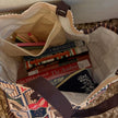 A close-up overhead view of a canvas tote bag filled with books, pencils, and a small zip pouch. The tote has a cream canvas body, dark brown handles, and a multicolor printed pattern on the exterior.