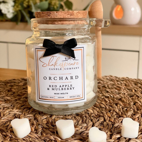 A glass jar candle from Shakespeare Candle Company in the Orchard scent, filled with white wax and topped with a cork lid. A black ribbon bow is tied around the neck, and a small wooden spoon rests on the side.
