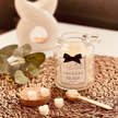 A glass jar candle labeled 'Shakespeare Candle Company Orchard' with a black bow graphic on the label, styled on a woven brown mat with eucalyptus leaves, wax cubes, and a wooden spoon.