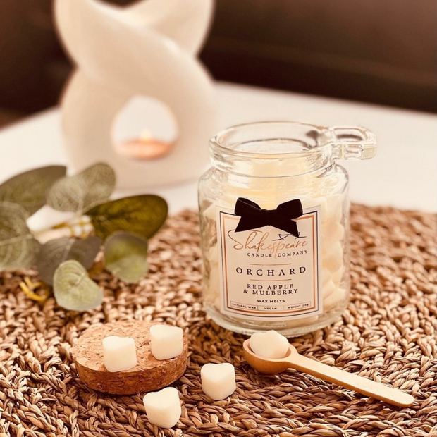 A glass jar candle labeled 'Shakespeare Candle Company Orchard' with a black bow graphic on the label, styled on a woven brown mat with eucalyptus leaves, wax cubes, and a wooden spoon.