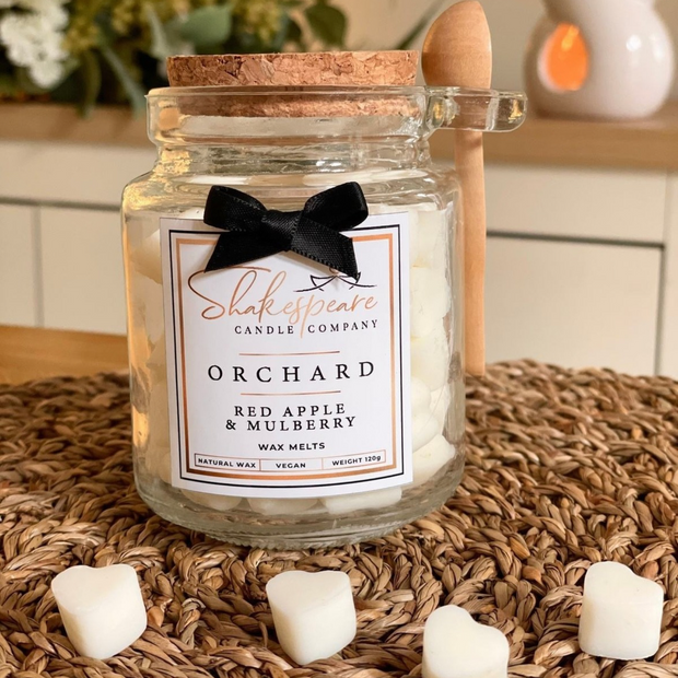 A glass jar candle from Shakespeare Candle Company in the Orchard scent, filled with white wax and topped with a cork lid. A black ribbon bow is tied around the neck, and a small wooden spoon rests on the side.