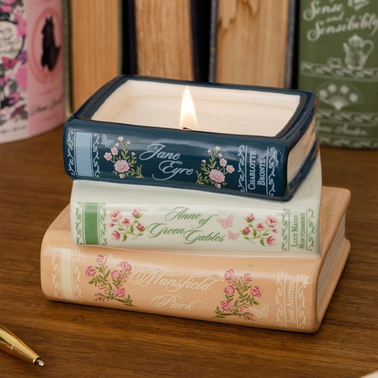 Three stacked ceramic book-shaped candle holders on a wooden surface, with the top one lit. The books feature floral spine designs and titles including Jane Eyre, Anne of Green Gables, and Mansfield Park.