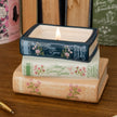 Three stacked ceramic book-shaped candle holders on a wooden surface, with the top one lit. The books feature floral spine designs and titles including Jane Eyre, Anne of Green Gables, and Mansfield Park.