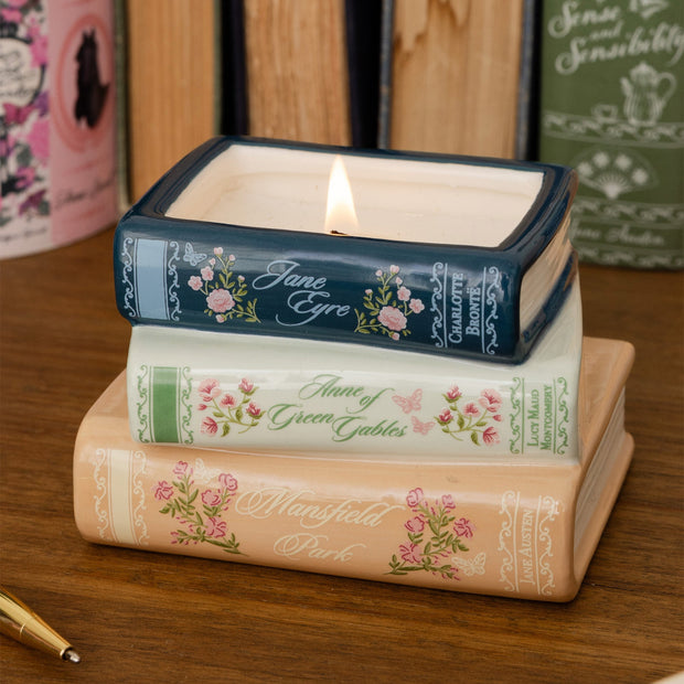 Three stacked ceramic book-shaped candle holders on a wooden surface, with the top one lit. The books feature floral spine designs and titles including Jane Eyre, Anne of Green Gables, and Mansfield Park.