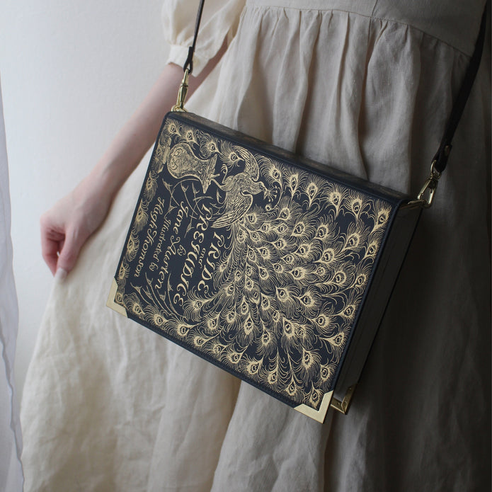 A person wearing a cream-colored dress carries a rectangular black shoulder bag with ornate gold peacock feather and script artwork, featuring gold corner protectors and a thin black strap.