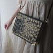 A person wearing a cream-colored dress carries a rectangular black shoulder bag with ornate gold peacock feather and script artwork, featuring gold corner protectors and a thin black strap.