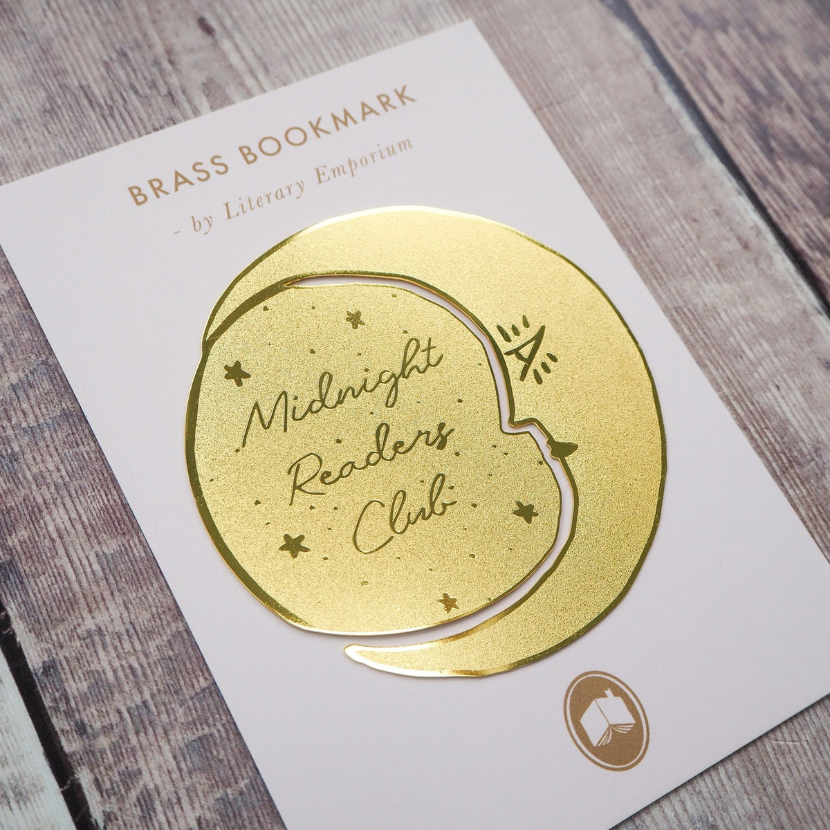 Gold brass bookmark on a white card with 'Midnight Readers Club' text, on a wooden surface.