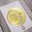 Gold brass bookmark on a white card with 'Midnight Readers Club' text, on a wooden surface.