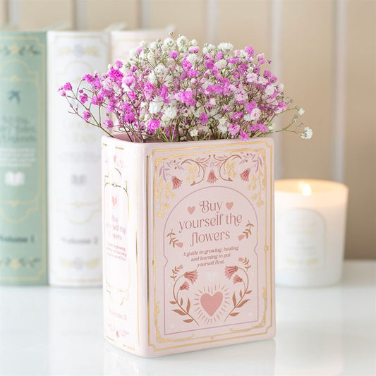 Decorative book shaped vase with pink and white flowers on a light surface with books and a candle in the background.
