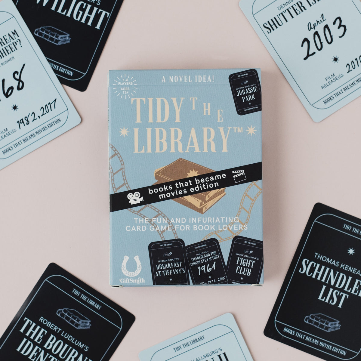 The book cover for 'Tidy the Library' card game is centered on a pale pink background, surrounded by scattered matching cards in black and light blue. The box shows the title in large cream text, a black banner reading 'books that became movies edition,' and several illustrated movie ticket graphics.