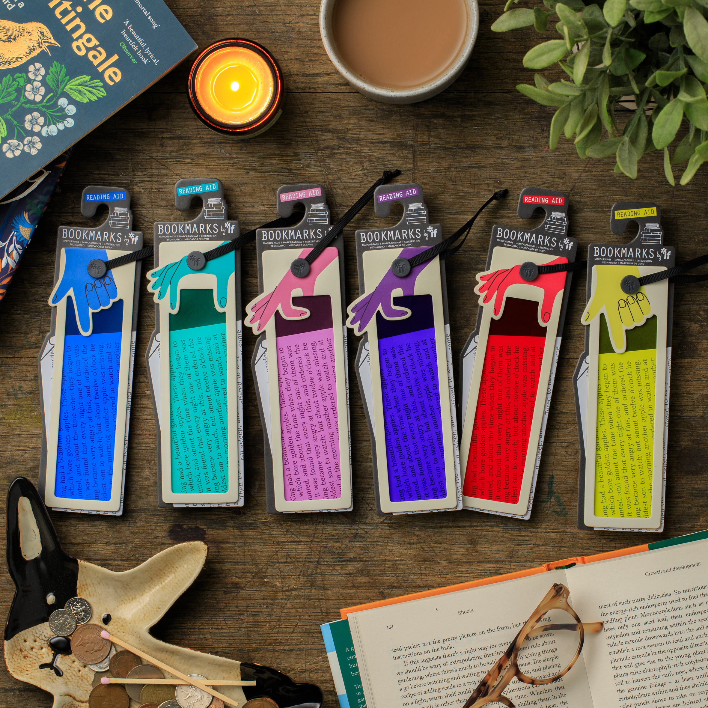 Six packaged paper bookmarks with colorful hand-shaped tabs are arranged on a wooden tabletop with books, a candle, a cup of tea, and a plant around them. The bookmarks come in blue, teal, pink, purple, red, and yellow colorways, each with text printed on the tab.