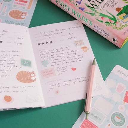 Open notebook with handwritten text and stickers on a green surface, with a book titled 'Emily Wildes' in the background.