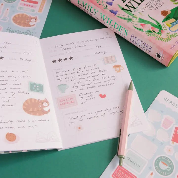 Open notebook with handwritten text and stickers on a green surface, with a book titled 'Emily Wildes' in the background.