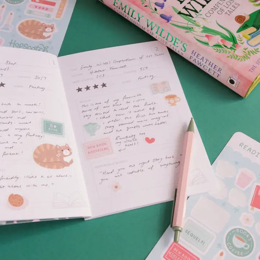 Open notebook with handwritten text and stickers on a green surface, with a book titled 'Emily Wildes' in the background.