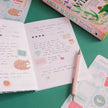 Open notebook with handwritten text and stickers on a green surface, with a book titled 'Emily Wildes' in the background.
