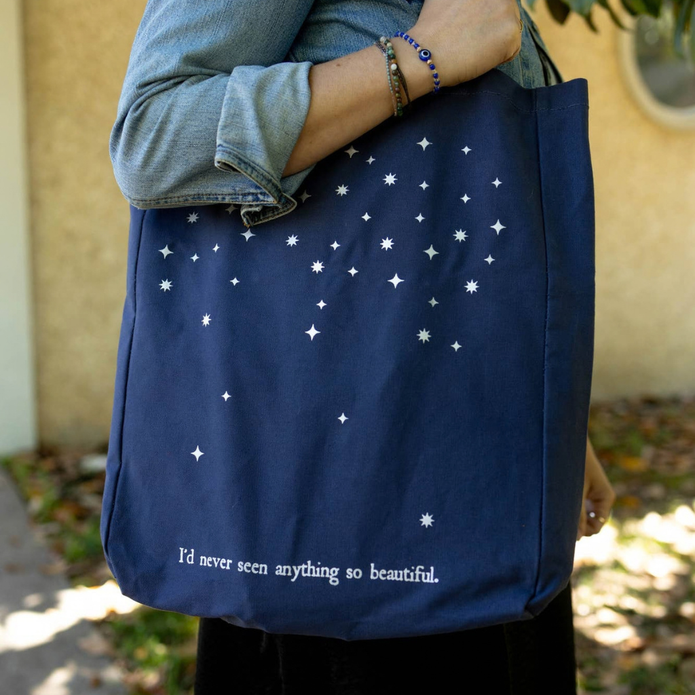 A navy blue canvas tote bag with white star and constellation prints and the text 'I'd never seen anything so beautiful.' at the bottom, carried by a person wearing a denim jacket outdoors.