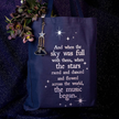 A navy blue canvas tote bag with white printed text and star graphics, featuring a hanging silver-toned star and boot charm. The bag is styled with dried flowers against a dark, textured, starry background.