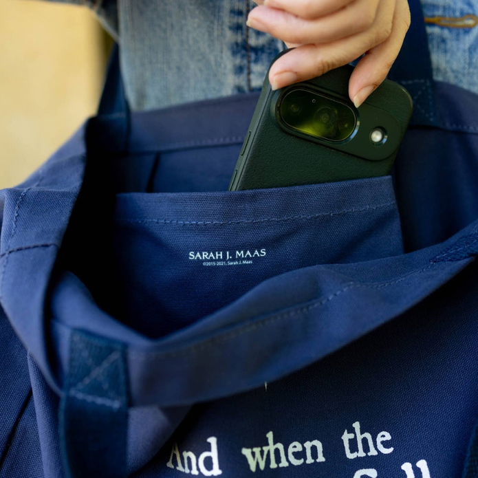 A navy blue fabric tote bag with a hand holding a black smartphone inside its side pocket. The brand name 'SARAH J. MAAS.